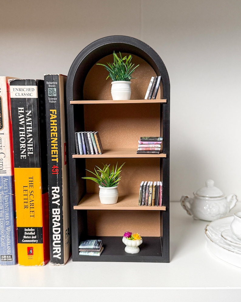 May include: A miniature black bookshelf with three shelves, featuring a selection of miniature books, potted plants, and a small flower arrangement. The bookshelf has an arched top and is made of wood.
