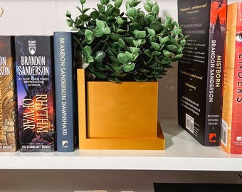 May include: A set of gold bookends with a potted plant on top. The bookends are on a white shelf with a variety of books, including "Rhythm of War" and "Mistborn: The Original Trilogy".