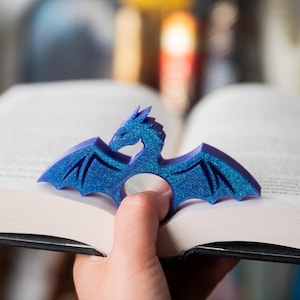 Könnte beinhalten: Ein blaues Drachen-Lesezeichen mit glitzernden Akzenten. Der Drache hat ausgebreitete Flügel und liegt auf den Seiten eines offenen Buches. Das Lesezeichen besteht aus einem festen Material und dient als Seitenmarkierung.