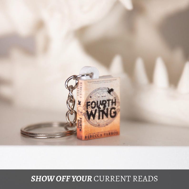 May include: A miniature book keychain featuring the title "Fourth Wing" by Rebecca Yarros. The book has an orange and tan cover with a circular design and a black bird. The keychain includes a silver ring and chain. The text "SHOW OFF YOUR CURRENT READS" is at the bottom.