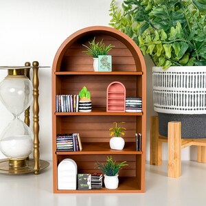 以下が含まれることがあります： アーチ型のトップが付いたミニチュア木製本棚。小さな本、装飾的な植物、小さなピンクの棚でいっぱいです。本棚は茶色で、3つの棚があります。植物は小さな鉢に入っています。背景には装飾的な砂時計があります。