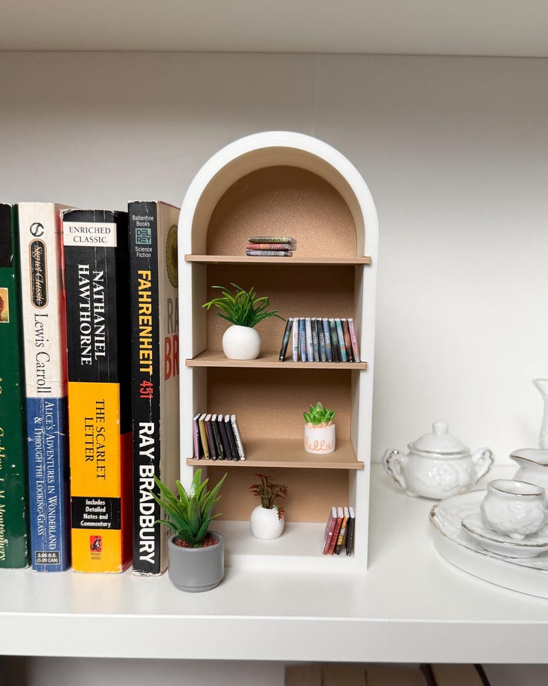 May include: A white miniature bookshelf with an arched top, featuring three shelves filled with miniature books, potted plants, and decorative items. The bookshelf is on a white shelf with a row of books behind it.