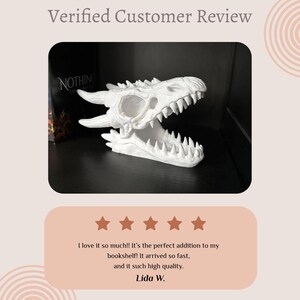 May include: A white dragon skull sculpture with sharp teeth and horns. The sculpture is displayed on a shelf with a book visible in the background. The image includes a customer review with five stars.