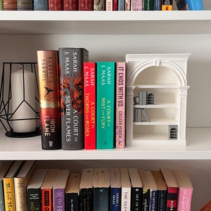 May include: A white bookshelf displays a collection of books, including titles by Sarah J. Maas and Colleen Hoover. A decorative miniature bookshelf and a black geometric candle holder are also present.