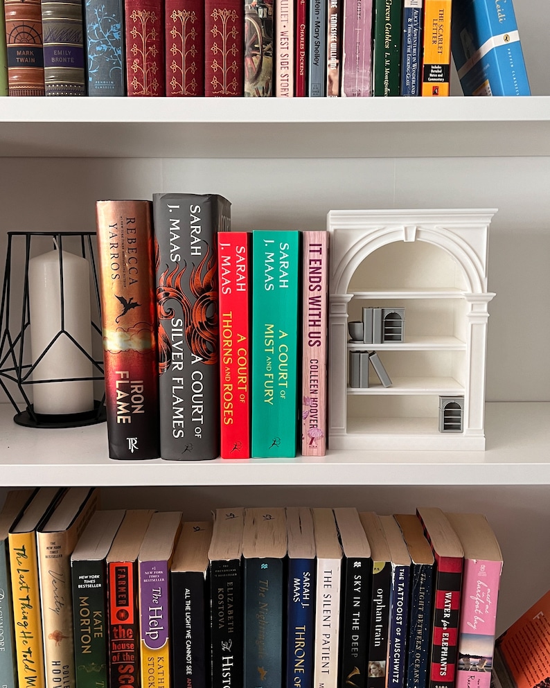 May include: A white bookshelf displays a collection of books, including titles by Sarah J. Maas and Colleen Hoover. A decorative miniature bookshelf and a black geometric candle holder are also present.