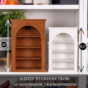 May include: Two miniature bookshelves are shown, one brown and one white. The brown bookshelf is 25.4 cm tall, and the white one is 21.6 cm tall. Both have an arched top and three shelves. Text at the bottom reads "2 SIZES TO CHOOSE FROM: 10 INCH VERSION | 8.5 INCH VERSION."