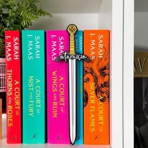 Könnte beinhalten: Ein schwertförmiger Lesezeichen befindet sich zwischen farbenfrohen Büchern. Die Bücher tragen Titel wie "A Court of Thorns and Roses" und "A Court of Silver Flames" von Sarah J. Maas. Auf dem Lesezeichen steht das Wort "ataraxia".
