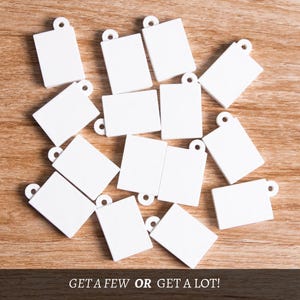 May include: A selection of small, white rectangular tags with a circular hole at the top, arranged on a wooden surface. The tags are plain, possibly for personalisation or labelling. The text at the bottom reads "GET A FEW OR GET A LOT!"
