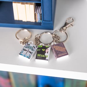 May include: Three miniature book keyrings, each depicting a book cover: "Quick Silver", "Wicked", and "Wuthering Heights". The keyrings are silver-toned and displayed on a white surface, with a blue bookshelf visible in the background.