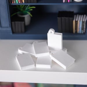 May include: A cluster of miniature white books, each with a small hole at the top, scattered on a white surface. These small, rectangular books are likely for a dollhouse or model. A blue bookshelf filled with books is visible in the background.