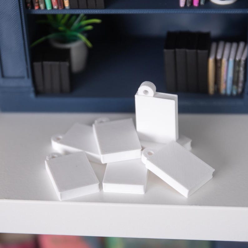 May include: A cluster of miniature white books, each with a small hole at the top, scattered on a white surface. These small, rectangular books are likely for a dollhouse or model. A blue bookshelf filled with books is visible in the background.