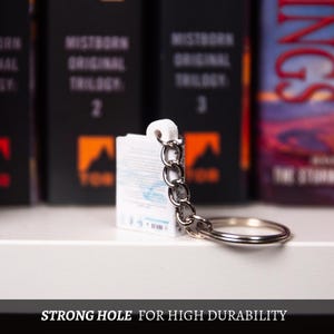 May include: A white keychain with a silver chain and ring. The keychain is attached to a small, white rectangular object. The text "STRONG HOLE FOR HIGH DURABILITY" is at the bottom of the image. Books are in the background.