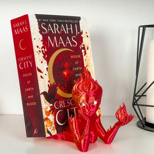 May include: A red 3D-printed figurine of a woman with flame-like hair, resting her chin on her hand, holding a flame. A book titled "Crescent City" by Sarah J. Maas is behind the figurine.