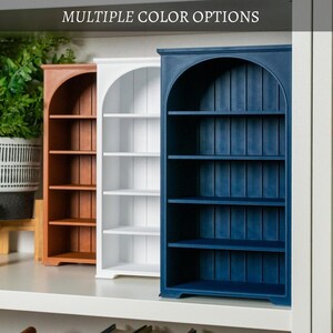 以下が含まれることがあります： 茶色、白、紺色の3色の木製本棚。 各本棚はアーチ型のトップと4つの棚があります。 テキスト「MULTIPLE COLOR OPTIONS」が棚の上に表示されています。