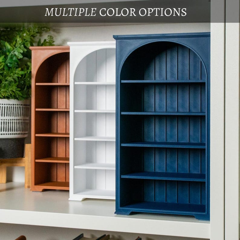 以下が含まれることがあります： 茶色、白、紺色の3色の木製本棚。 各本棚はアーチ型のトップと4つの棚があります。 テキスト「MULTIPLE COLOR OPTIONS」が棚の上に表示されています。