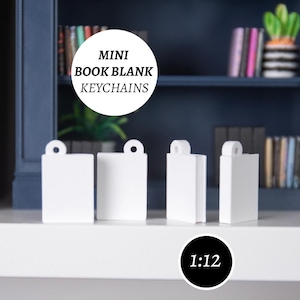 Może przedstawiać: Cztery białe mini breloczki do książek. Każdy brelok ma prostokątny kształt z małą pętlą na górze. Breloki są wyświetlane na białej powierzchni z ciemnoniebieskim tłem. Tekst "MINI BOOK BLANK KEYCHAINS" znajduje się w białym okręgu.