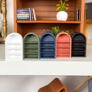 Puede incluir: Cinco estanterías arqueadas en miniatura en blanco, verde, azul, coral y negro. Cada estantería tiene tres niveles. Las estanterías están dispuestas sobre una superficie blanca, con una estantería de madera y objetos decorativos en el fondo.