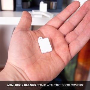 May include: A tiny, white, blank book model with a hole at the top, held in a hand. The text at the bottom of the image reads, "MINI BOOK BLANKS COME WITHOUT BOOK COVERS."