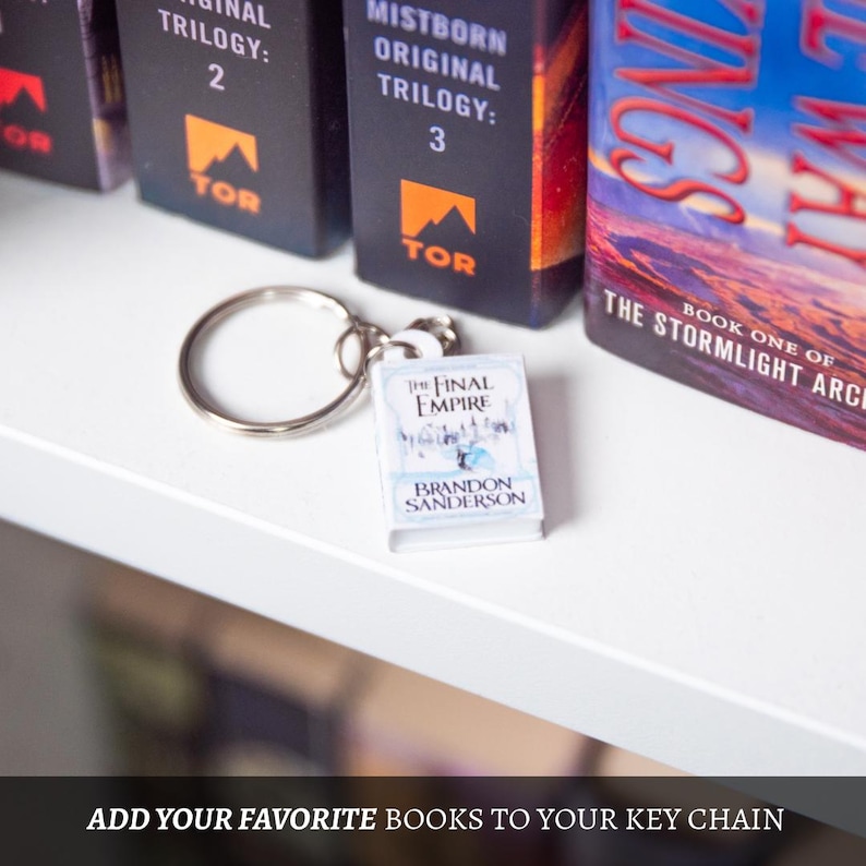 May include: A silver keychain with a miniature book charm. The book charm is titled "The Final Empire" by Brandon Sanderson. The keychain is on a white shelf, with book spines in the background.