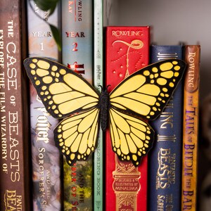 May include: A decorative butterfly-shaped object with bright yellow wings and black vein patterns, positioned horizontally between several hardcover books. The butterfly features a textured black central body and symmetrical wing designs.