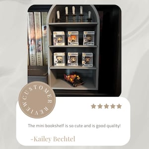 以下が含まれることがあります： 3段の棚がある灰色のミニチュア本棚。箱に入ったいくつかのFunko Popフィギュアと小さな玩具列車が展示されています。棚の上部には装飾的な燭台があります。左側には本が見えます。テキストには「The mini bookshelf is so cute and is good quality!」と書かれています。