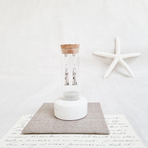 Puede incluir: Un vial de vidrio transparente con un tapón de corcho, que contiene pendientes de faro plateados. El vial descansa sobre una base de cerámica blanca y un cuadrado de tela gris. Una estrella de mar blanca está en el fondo.