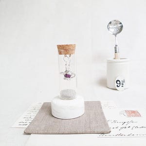 Könnte beinhalten: Silberne Teekannen-Ohrringe mit einem violetten Edelstein, ausgestellt in einem Glasfläschchen mit einem Korkverschluss, auf einem weißen Marmorsockel. Das Fläschchen ist mit silbernen Perlen gefüllt.