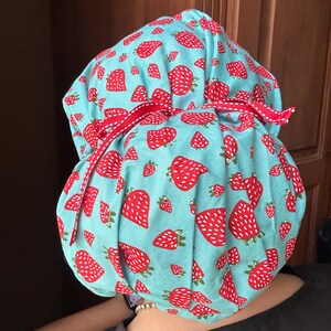 Puede incluir: Un gorro de quir&oacute;fano azul turquesa con estampado de fresas blancas y rojas. El gorro tiene la parte superior fruncida y est&aacute; atado con una cinta roja.