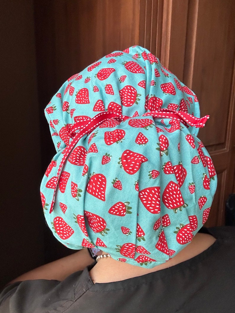 Puede incluir: Un gorro de quir&oacute;fano azul turquesa con estampado de fresas blancas y rojas. El gorro tiene la parte superior fruncida y est&aacute; atado con una cinta roja.