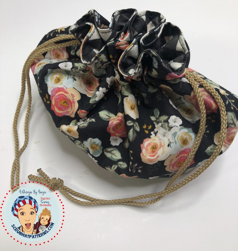 May include: A black drawstring bag with a floral print featuring pink, white, and yellow roses. The bag has a tan drawstring and is made of fabric.