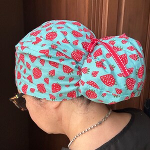 Puede incluir: Un gorro de quir&oacute;fano turquesa con estampado de fresas y un lazo rojo. El gorro est&aacute; atado en un mo&ntilde;o en la parte posterior.