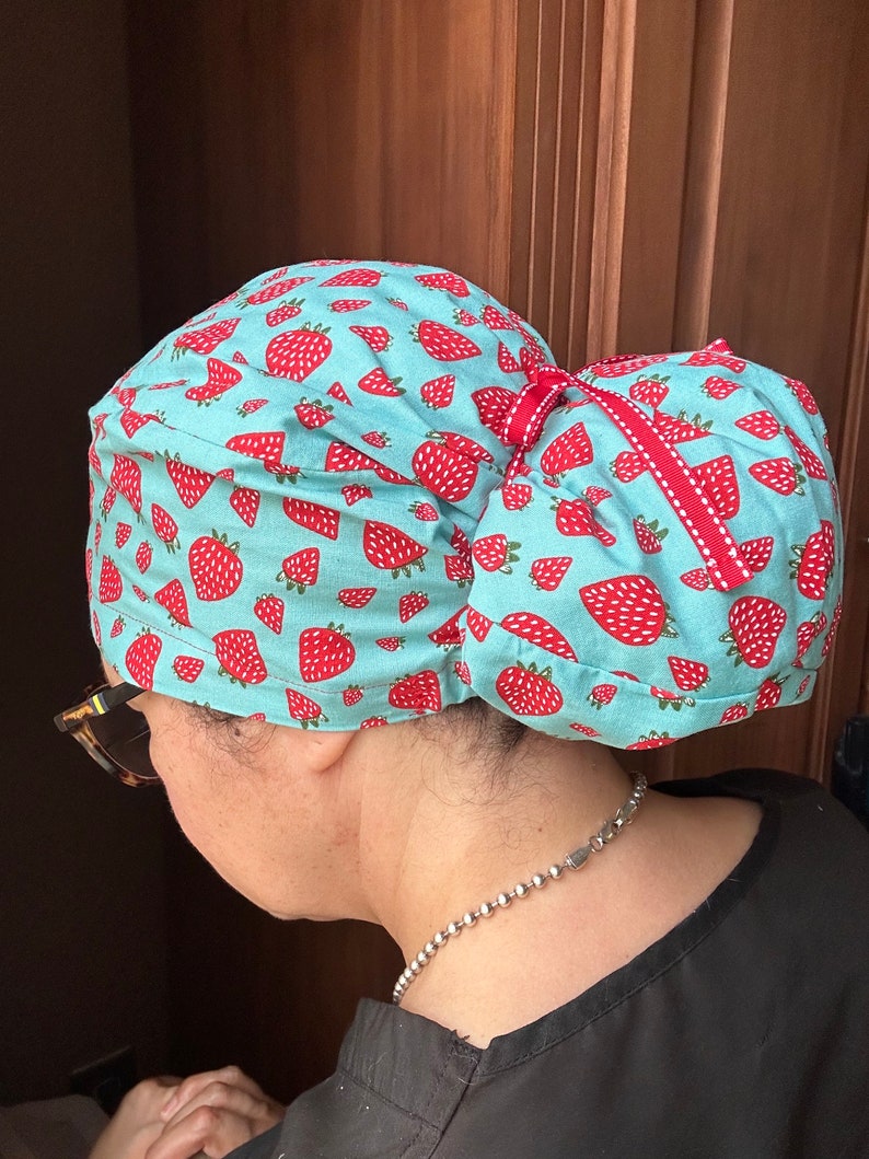 Puede incluir: Un gorro de quir&oacute;fano turquesa con estampado de fresas y un lazo rojo. El gorro est&aacute; atado en un mo&ntilde;o en la parte posterior.