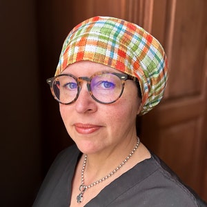 May include: A person wearing a black scrub top with a silver chain necklace and a plaid fabric head covering. The person is wearing glasses and has a neutral expression.