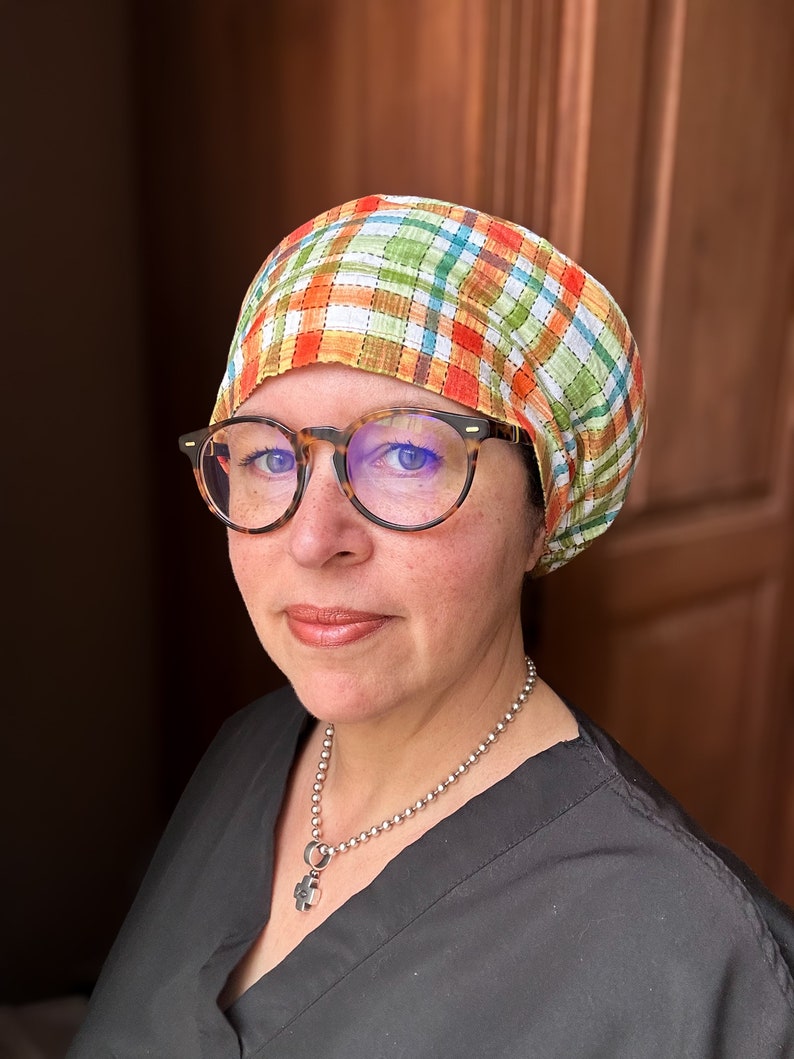 May include: A person wearing a black scrub top with a silver chain necklace and a plaid fabric head covering. The person is wearing glasses and has a neutral expression.