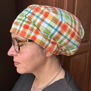May include: A person wearing a green, orange, and brown plaid scrub cap with a silver chain necklace. The cap is tied at the back of the head.