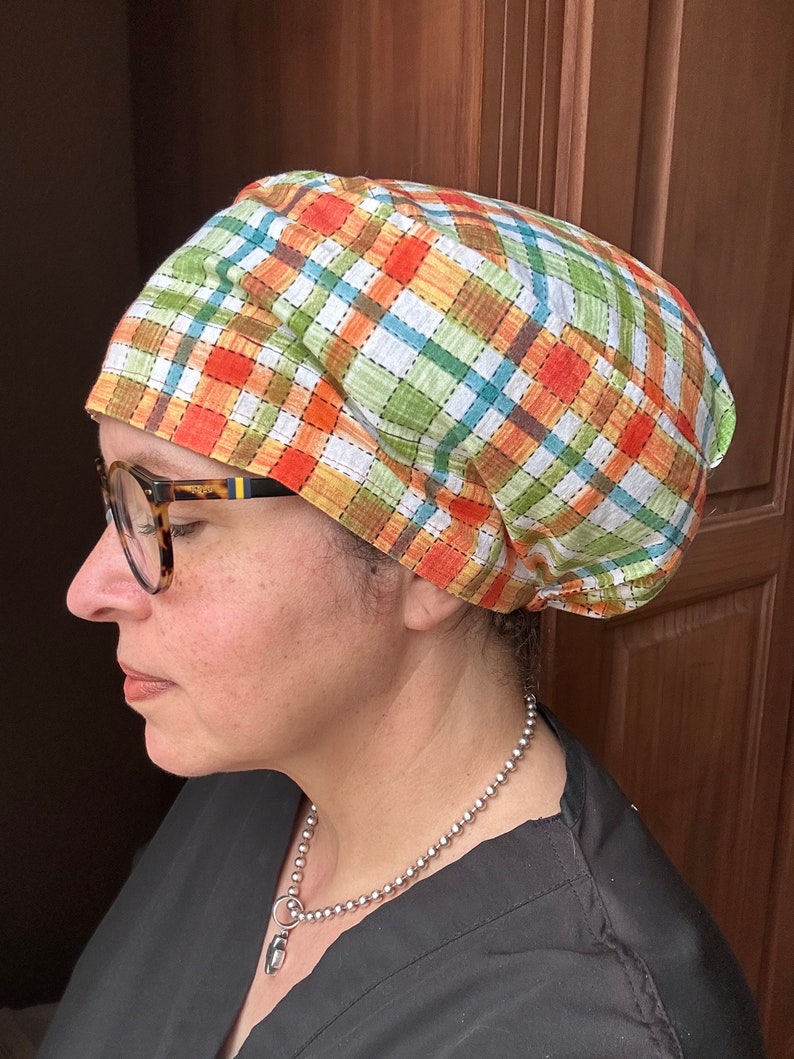 May include: A person wearing a green, orange, and brown plaid scrub cap with a silver chain necklace. The cap is tied at the back of the head.