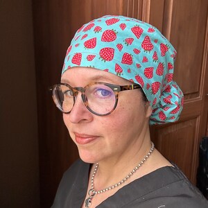Puede incluir: Una mujer con una cofia quir&uacute;rgica azul claro con un patr&oacute;n de fresas rojas. Tambi&eacute;n lleva una bata quir&uacute;rgica negra con un collar de cadena plateada.