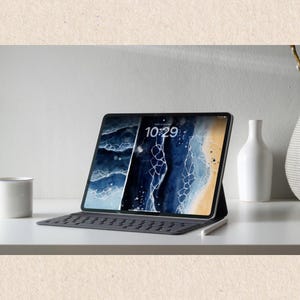 Puede incluir: Una tableta negra con funda de teclado y l&aacute;piz &oacute;ptico, que muestra una imagen abstracta azul y dorada en la pantalla. La hora 10:29 es visible. Una taza y un jarr&oacute;n blancos est&aacute;n sobre la superficie blanca.