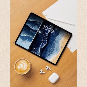 Puede incluir: Una tableta digital que muestra un dise&ntilde;o abstracto azul y blanco, con la hora 10:29 en la pantalla. Una taza de caf&eacute;, auriculares inal&aacute;mbricos y un estuche de carga est&aacute;n colocados sobre una superficie de madera, junto con un l&aacute;piz &oacute;ptico blanco y una hoja de papel.