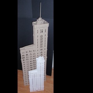 May include: Two architectural models of skyscrapers. The larger model is beige with a tall, rectangular design and a spire. The smaller model is white and has a similar design. Both models have numerous windows.