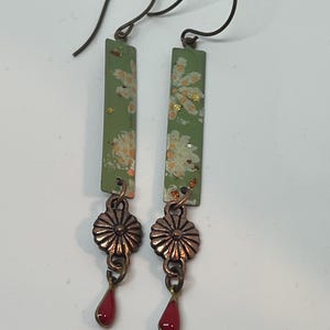 May include: A pair of green dangle earrings with a floral pattern, copper flower accents, and red teardrop dangles.