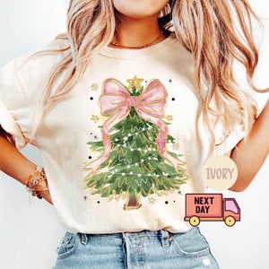 Pode incluir: Uma camiseta branca com uma ilustração em aquarela de uma árvore de Natal com um laço rosa e luzes douradas. O texto "IVORY" e "NEXT DAY" é visível na imagem.