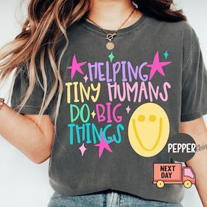 May include: A charcoal gray short-sleeve t-shirt featuring the text HELPING TINY HUMANS DO BIG THINGS in purple, yellow, pink, and teal lettering. The design includes pink stars, small teal and pink sparkles, and a large yellow smiley face.