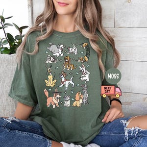 May include: Moss green t-shirt featuring a colorful cartoon dog graphic. The graphic includes various dog breeds and small blue hearts. The shirt is a relaxed fit and is being worn by a person.