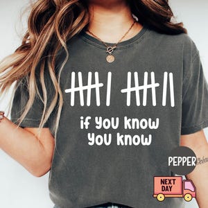 May include: Dark gray t-shirt with white graphic text and tally marks. The text reads "if you know you know". The shirt has short sleeves and a crew neck. A small pink delivery truck graphic is visible.