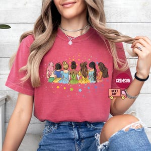 Puede incluir: Una camiseta roja con un gr&aacute;fico de siete mujeres de dibujos animados con cabello largo y vestidos coloridos. Las mujeres est&aacute;n de pie en una fila, mirando al espectador. El gr&aacute;fico est&aacute; rodeado de estrellas.