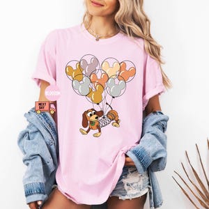 May include: A pink t-shirt with a cartoon image of a dog holding onto a bunch of colorful balloons shaped like Mickey Mouse ears. The dog is wearing a brown vest and has a spring attached to its body.