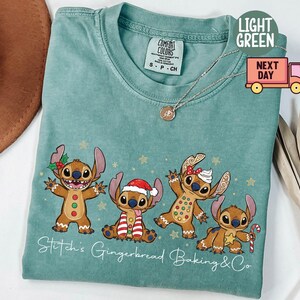 Puede incluir: Camiseta Comfort Colors verde claro con un dise&ntilde;o festivo de cuatro personajes de dibujos animados disfrazados de hombrecitos de jengibre. El texto "Stitch's Gingerbread Baking & Co" est&aacute; impreso debajo de los personajes. Un collar dorado est&aacute; sobre la camiseta.