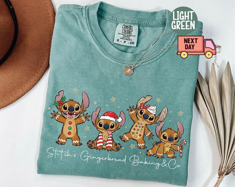 Puede incluir: Camiseta Comfort Colors verde claro con un dise&ntilde;o festivo de cuatro personajes de dibujos animados disfrazados de hombrecitos de jengibre. El texto "Stitch's Gingerbread Baking & Co" est&aacute; impreso debajo de los personajes. Un collar dorado est&aacute; sobre la camiseta.