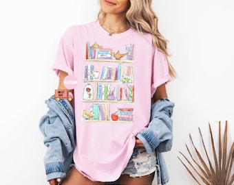 May include: Pink t-shirt featuring a watercolor illustration of a bookshelf filled with books, a rose, a teapot, and an apple. The shirt has short sleeves and a relaxed fit. The word "Blossom" is printed on a pink circle.
